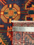 Close-up of a patterned rug with a stainless steel ruler placed vertically to show the pile height and weave detail. The rug features a dark navy blue base with geometric medallions in rust red, beige, orange, and green.