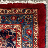 A close-up corner of a patterned area rug on a gray carpet. The rug has a deep red border, cream and blue inner fields, and white fringe.