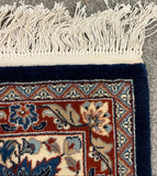 Close-up of a patterned area rug corner with off-white fringe, showing a navy blue border and red, cream, and blue floral field on a gray carpet background.