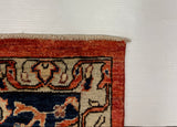 A close-up corner view of a patterned rug showing a rust red border, cream field, and dark navy sections with stylized motifs.