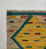Close-up corner of a woven rug with a geometric pattern in yellow, teal, pink, red, and beige, laid on a white surface.